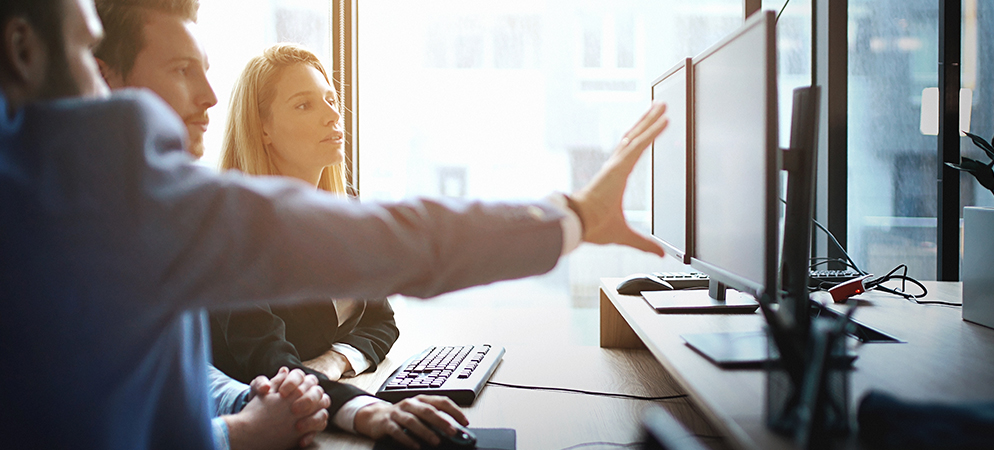 Team reviewing data on dual monitors in modern office, coworkers collaborating at desktop workstation with sunlight and business analytics focus
