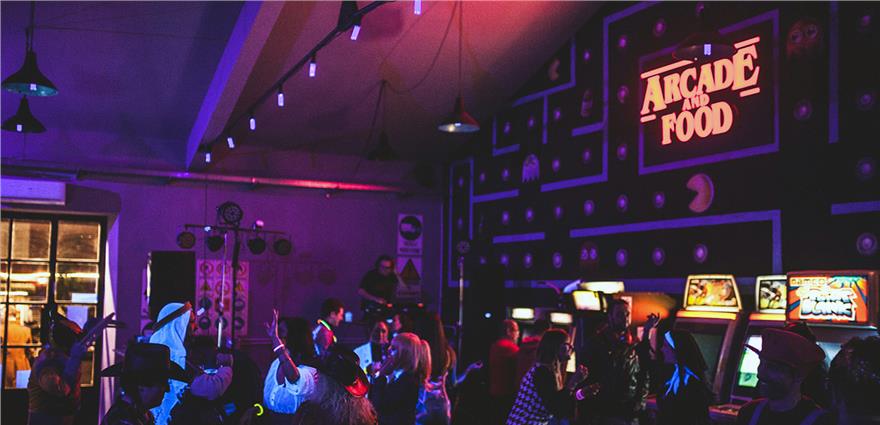 Crowded retro arcade bar with neon “Arcade and Food” sign, classic machines and people partying under colorful lights.