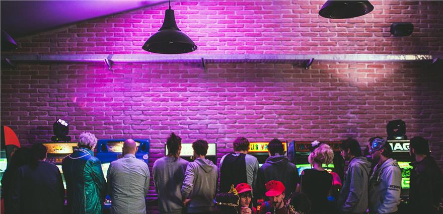 Players lined up at classic arcade machines in a retro bar with exposed brick walls and purple lighting.