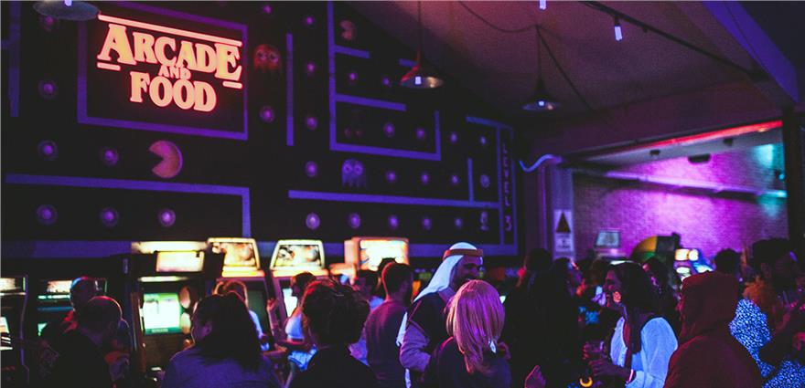 Crowded retro arcade bar with neon “Arcade and Food” sign, classic game machines and people partying under colorful lights.