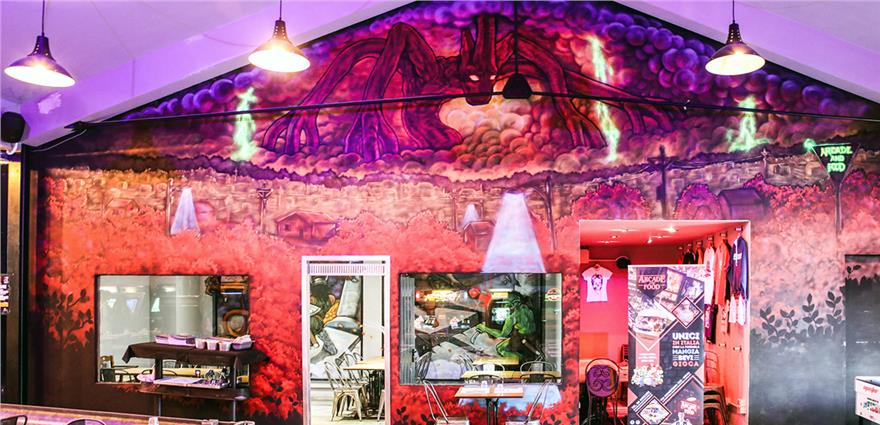 Colorful fantasy-themed mural with dragon artwork inside an arcade bar, above food counter and merchandise area.