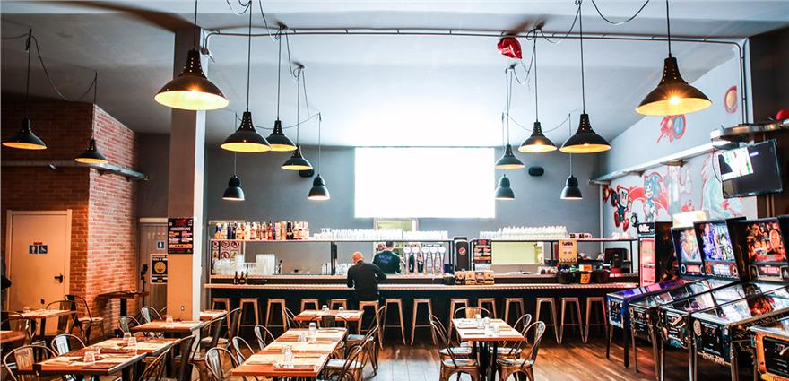 Arcade bar with long counter, hanging industrial lights, rows of pinball machines and tables set for dining.