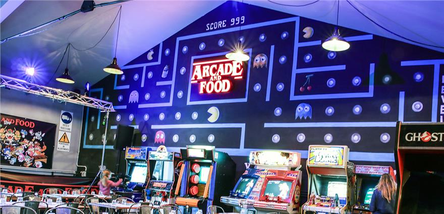 Retro arcade bar with Pac‑Man themed wall mural, neon “Arcade and Food” sign and rows of classic arcade machines.