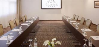 Hotel Aristos meeting room with two rows of tables facing a screen, set with water bottles and notepads.
