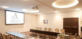 Hotel meeting room arranged in a U-shape with tables, chairs, water bottles, and a projection screen displaying “Hotel Aristos.”