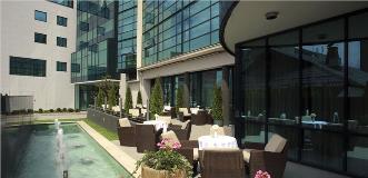 Modern hotel courtyard with wicker chairs and round tables beside a narrow fountain, bordered by glass-walled buildings.