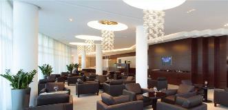 Elegant hotel lounge with clusters of brown leather armchairs, modern chandeliers, and a wood-paneled wall with a TV.