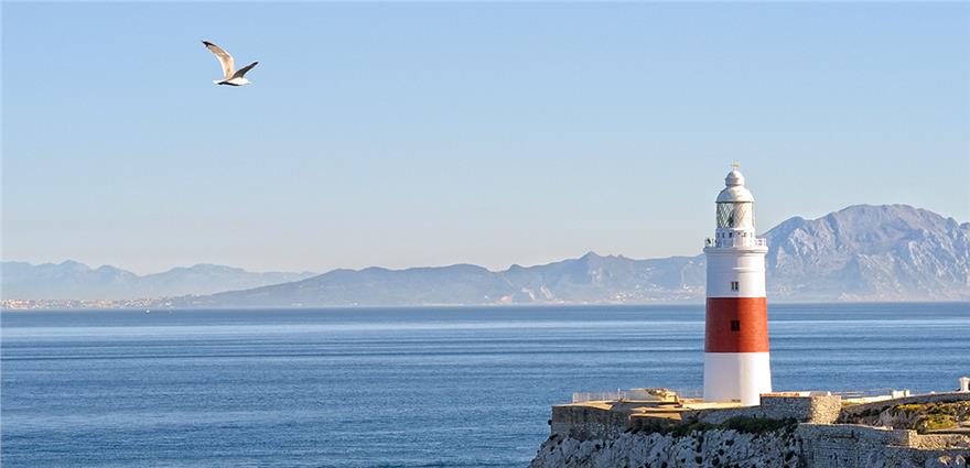 Coastal lighthouse with red stripe on rocky point overlooking calm blue sea and distant mountains, seagull flying above.