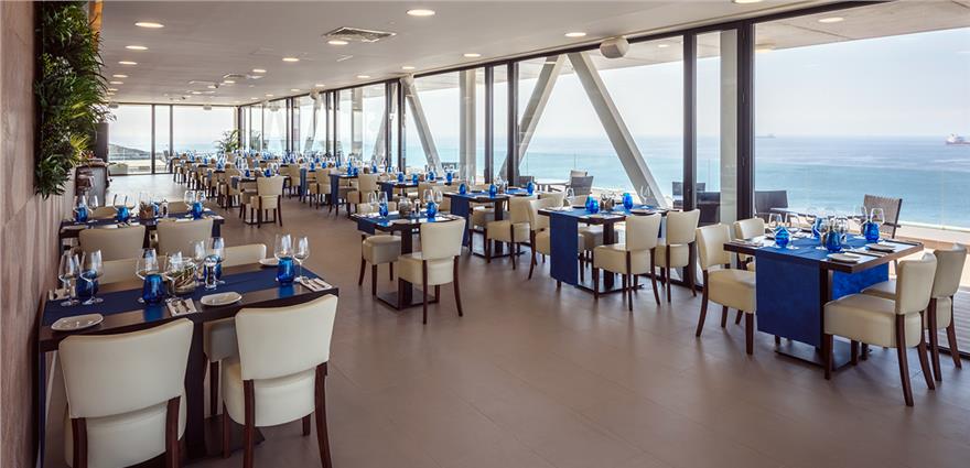 Spacious ocean-view restaurant with floor‑to‑ceiling windows, cream chairs and blue table runners overlooking the sea.