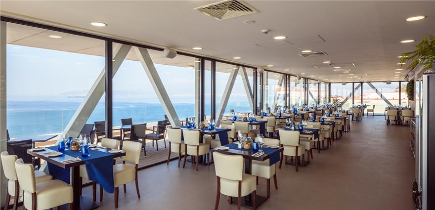 Elegant seaside restaurant with floor‑to‑ceiling windows, cream chairs, blue table settings and panoramic ocean views.