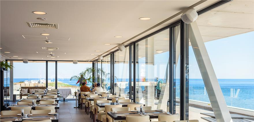 Bright seaside restaurant with floor‑to‑ceiling windows, cream chairs and panoramic ocean views.