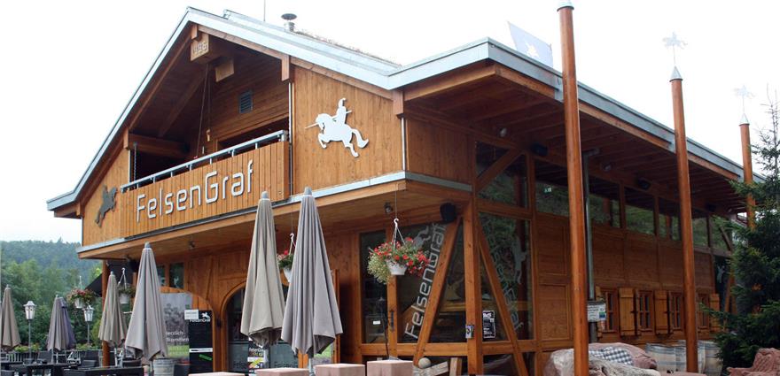 Wooden alpine-style restaurant with a balcony, signage reading "FelsenGraf," and outdoor seating.