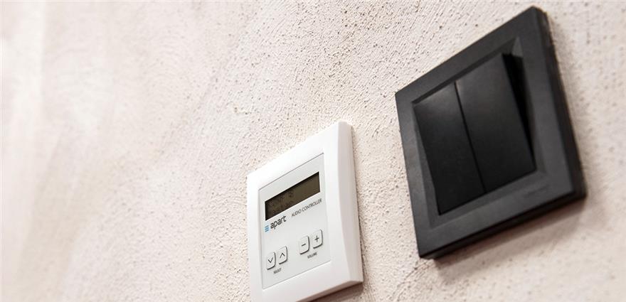 White digital audio control and black wall switch on a textured beige wall.