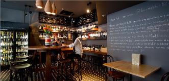 Wine bar interior with a chalkboard menu, wall of wine bottles, and a wooden bar with stools.