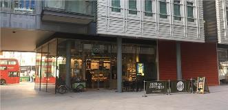 Modern cafe storefront with glass walls, outdoor seating, and a red bus outside.