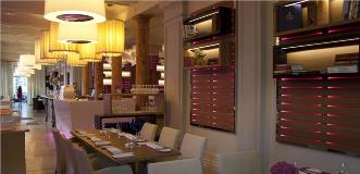 Modern restaurant interior with yellow pendant lights, wooden beams, and pink glow along wall shelves.