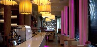 Contemporary restaurant interior with a long white bar, warm yellow pendant lights, and pink lighting accents.