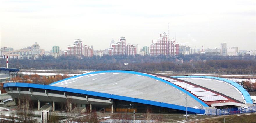 Low, dome-shaped sports arena with a blue trim roof in the foreground, with a city skyline of tall buildings behind it.