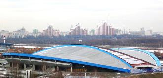 Large covered sports arena with curved blue roof set against a city skyline of high-rise buildings.