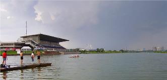 Rowing regatta venue with grandstands by the shore, people on docks, and a team rowing on a wide, calm course.