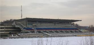 Large covered grandstand with red, blue, and gray seating overlooking a snow-covered rowing or water sports venue.