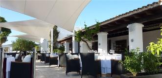 Elegant outdoor Mediterranean restaurant terrace with white sail shades, wicker chairs and tables set for fine dining.