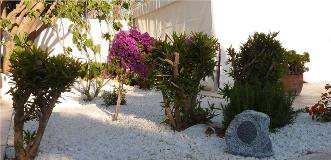 Mediterranean garden with white gravel, flowering shrubs and a rock-shaped outdoor speaker near a white wall.