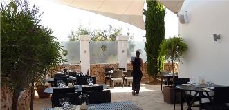 Elegant outdoor restaurant terrace with modern black tables, white sunshade, stone walls and greenery.