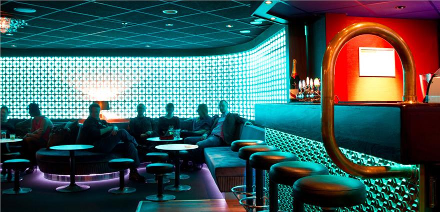 Neon-lit lounge bar with curved turquoise backlit wall, booth seating, and high stools at the counter.