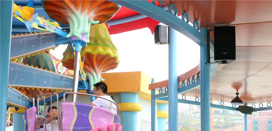 Colorful spinning amusement ride with children in pastel cars beneath a canopy with mounted speakers.
