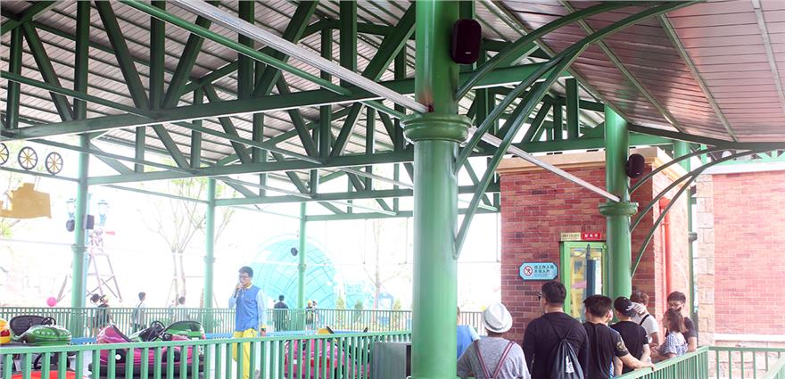 Covered queue area at an amusement park with green metal beams, brick wall, and people waiting in line.