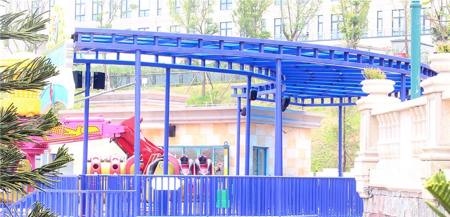 Brightly colored amusement park area with blue track structure and red ride seats behind a blue fence.