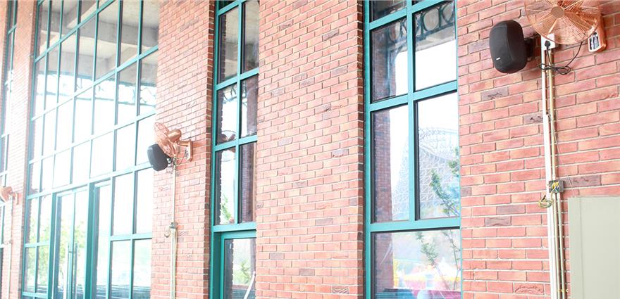 Brick building facade with large green-framed windows, wall-mounted speakers, and industrial fans.