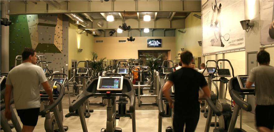 Busy gym with people using treadmills and ellipticals, rock-climbing wall in background, and ceiling-mounted speakers.