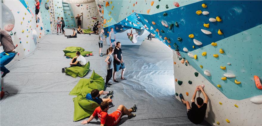 Indoor bouldering gym with climbers on colorful walls and others relaxing on green crash pads.