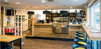 Cozy café with wooden counter, display case of pastries, bar stools, and large windows along one side.