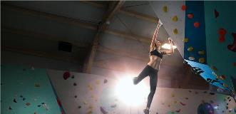 Woman bouldering on an overhanging indoor climbing wall, lit dramatically from below.