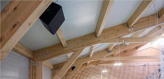 Ceiling of a sports hall with exposed wooden beams, hanging lights, and a mounted black speaker.