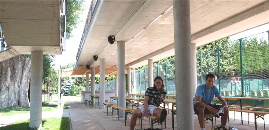 Two people sit on benches under a concrete overhang beside outdoor sports courts and green trees.