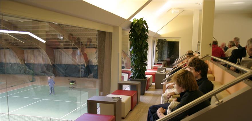 Spectators seated in a glass-enclosed lounge overlooking an indoor tennis court.