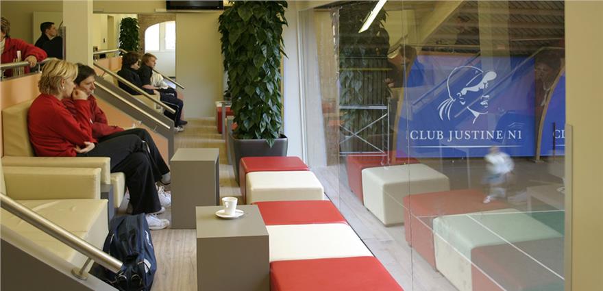 Spectators seated in a lounge overlooking indoor tennis courts at Club Justine N1 through large glass windows.