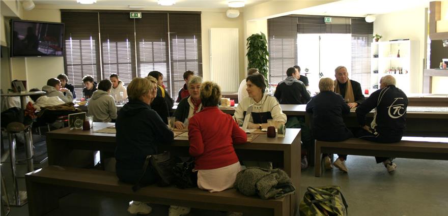 People socializing at long wooden tables inside a tennis club café or lounge area.
