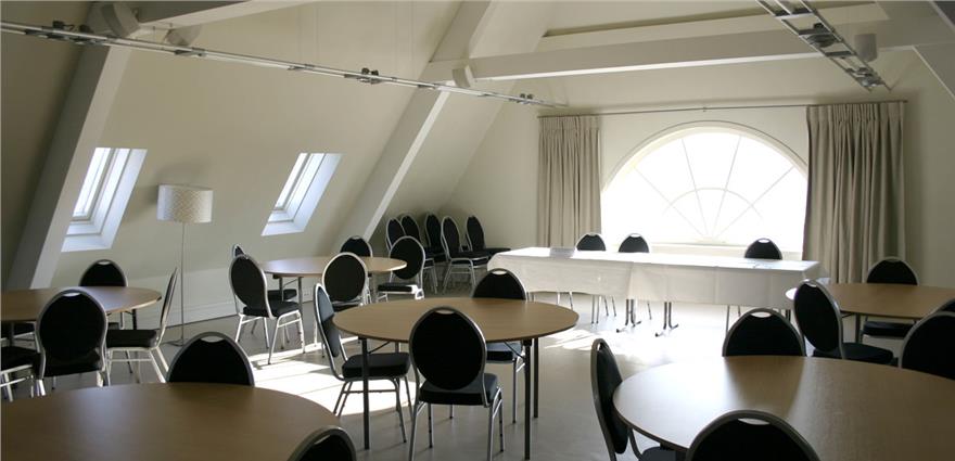 Bright conference or banquet room with round tables, black chairs, and a large arched window.