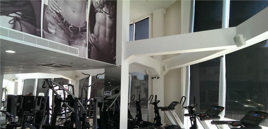 Bright gym interior with cardio machines by tall windows and large black‑and‑white fitness posters on the walls.