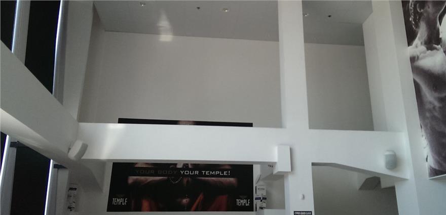 High-ceilinged gym interior with white beams, motivational poster reading “YOUR BODY YOUR TEMPLE!” and wall speakers.