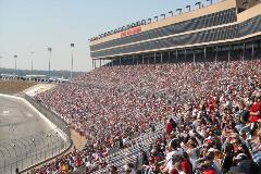 Crowded grandstands at a motor speedway overlooking a racetrack during a major racing event.