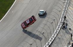 Red race car lifted onto two wheels after impact against the track wall, with another car and crew nearby.