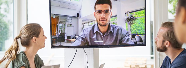Small team in meeting room video conferencing with remote colleague displayed on wall-mounted screen