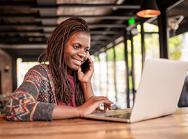 Woman working remotely in a café, smiling while talking on smartphone and using laptop