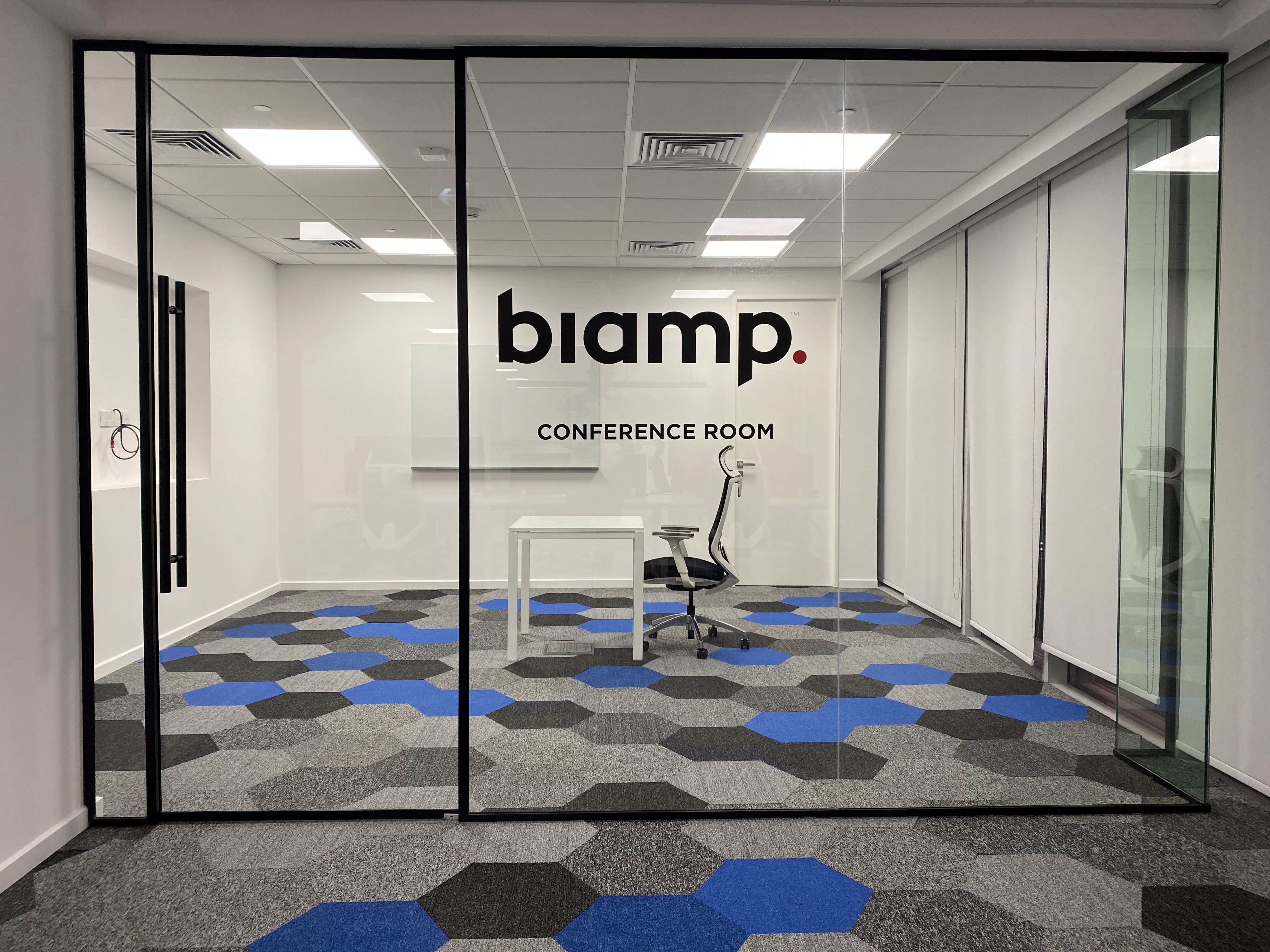 Modern Biamp conference room with glass walls, single table, office chair, and geometric blue‑gray carpet